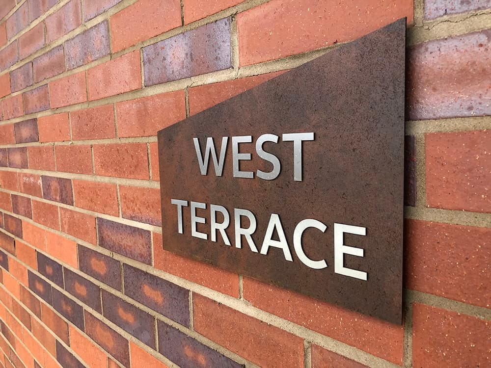 Close up of a sign identifying the West Terrace within Brook Place Apartments. Features stainless steel letters applied onto a mono pitch shaped sign made from a Corten steel laminate