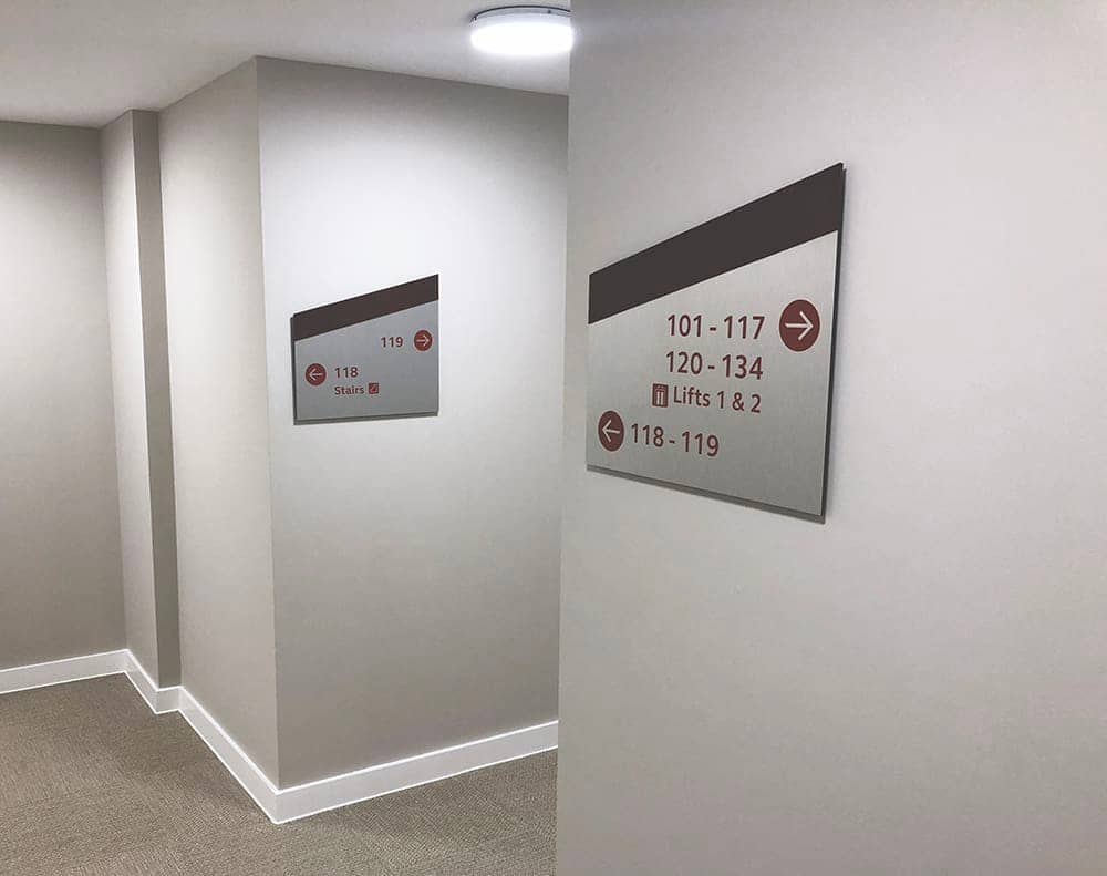 Internal direction signs within Brook Place Apartments, in a mono pitch shape with a dark brown band on the top and corten vinyl coloured letters applied on brushed stainless steel background