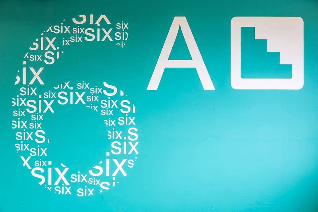 Large scale graphic applied to a wall in a car park showing a block of turquoise colour with a graphic in the shape of a number six, filled with the word six in white lettering of various sizes. Alongside is a capital A and the pictogram used to denote stairs, both in white.