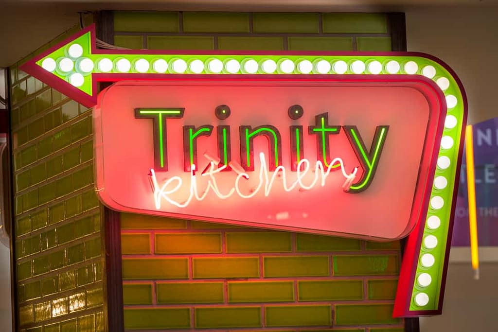 Large illuminated directional sign to Trinity Kitchen, attached to a column finished in green metro tiles. Trinity is written as individual letters, with green illumination with kitchen in a pink neon running across these at angle. The name is contained in a lozenge shape, with an arrow running along the outside from the rights edge across the top. The arrow has white spot lights running along a central recess on a green background. The sides of the arrow shape are pink.