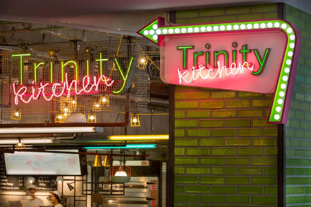 Stepped back view showing an identity sign and direction sign for Trinity Kitchen in situ. In both Trinity features as individual letters with green illumination running through the centre. The word kitchen is written in pink neon italics and runs at an angle. The identity sign is suspended from the ceiling and behind a wire mesh screen. The directional sign is attached to a green-tiled column within a lozenge shape. An arrow runs around the right and top edge of the lozenge which is illuminated by white inset spot lights