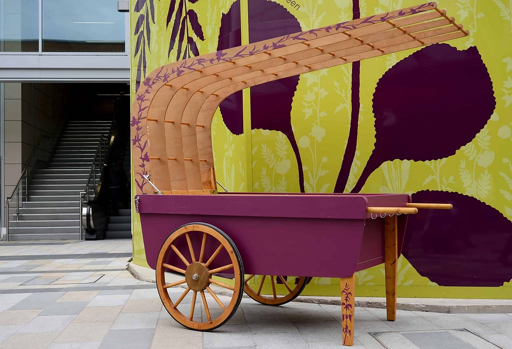 Community barrow, based on a traditional costermonger barrow with wheels and wooden overarching canopy. The base is painted in aubergine and the rest is left in natural wood with a painted leaf motif in aubergine snaking up the leg rests and the canopy edge.