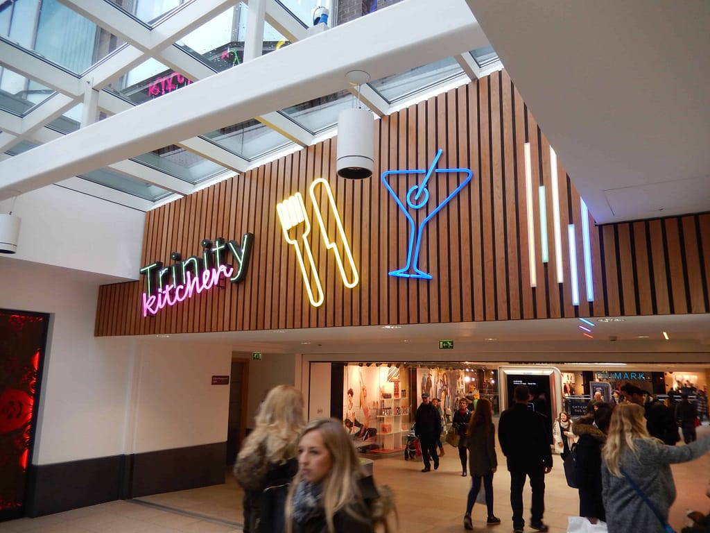 Neon signs applied to a wooden panelled background featuring the Trinity Kitchen identity; a large yellow outline of a knife and fork; a large blue outline of a cocktail glass with a cherry on a stick; and vertical lines of various lengths and colours