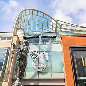 Large letter T in a gothic style, applied above the entrance to The Trinity Leeds shopping centre. The sign is formed from a stainless-steel outline and filled with individual metal discs to give it sparkle. In front of the sign is a metal mesh sculpture of the goddess Minerva.