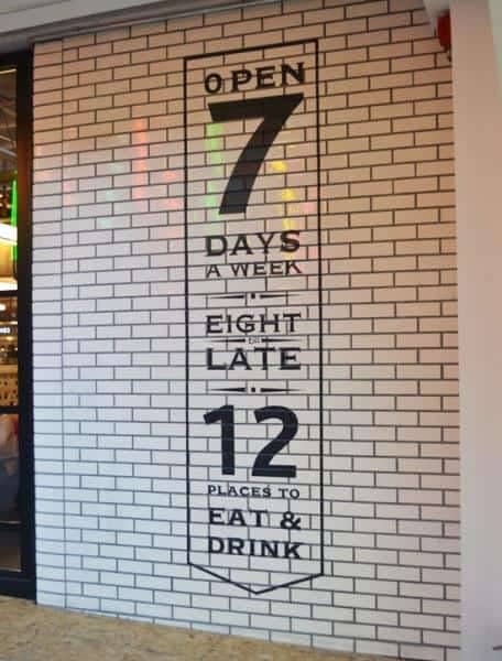 Decorative, floor to ceiling white tiled wall panel. Featuring the words Open 7 days a week, eight till late, 12 places to eat and drink in black lettering..