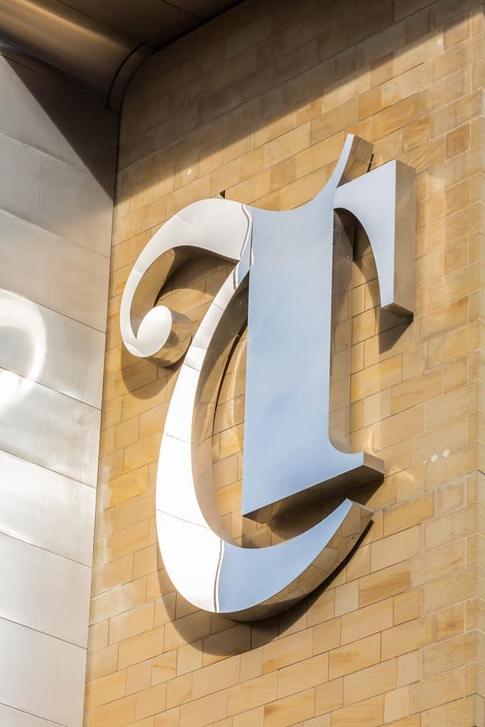Image of a large letter T in a gothic style made from polished stainless steel applied to a sandstone coloured brick wall.