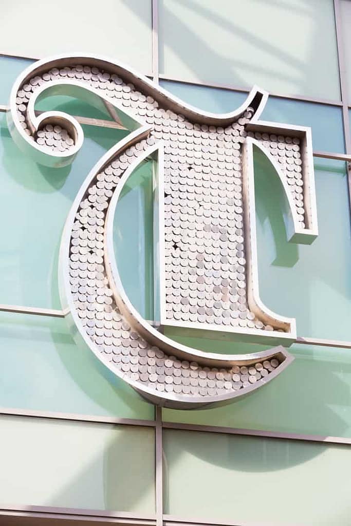 Close up of a the entrance identification sign for Trinity Leeds shopping centre. If feature the outline of a letter T, in a gothic style that is infilled with small metal discs
