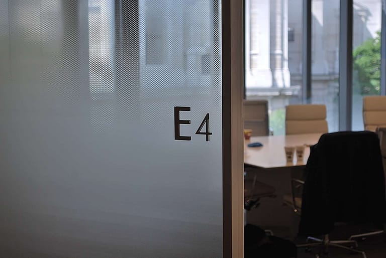 View through the open door to an office. The glazed office wall is covered in manifestation which has a diffused pattern design. The room name E4 is applied to the glass as individual characters