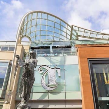 Large letter T in a gothic style, applied above the entrance to The Trinity Leeds shopping centre. The sign is formed from a stainless-steel outline and filled with individual metal discs to give it sparkle. In front of the sign is a metal mesh sculpture of the goddess Minerva.